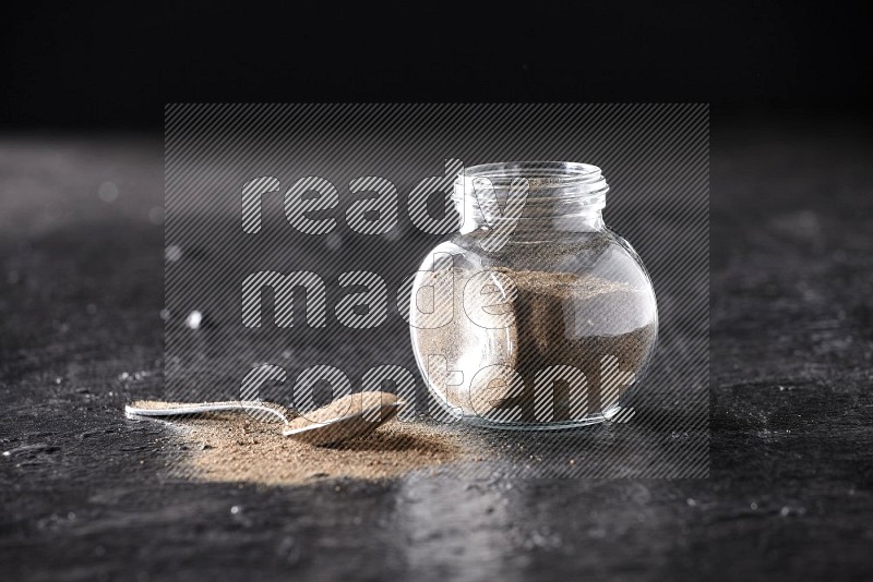 A glass spice jar full of black pepper powder and a metal spoon full of powder on textured black flooring