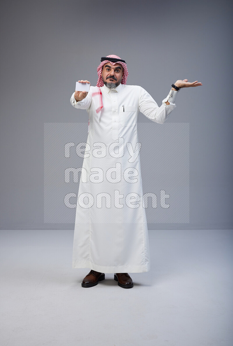 Saudi man Wearing Thob and red Shomag standing holding ATM card on Gray background