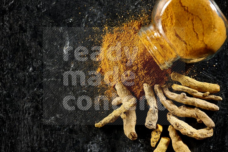 A flipped glass spice jar full of turmeric powder and powder spilled out of it with dried whole fingers on textured black flooring