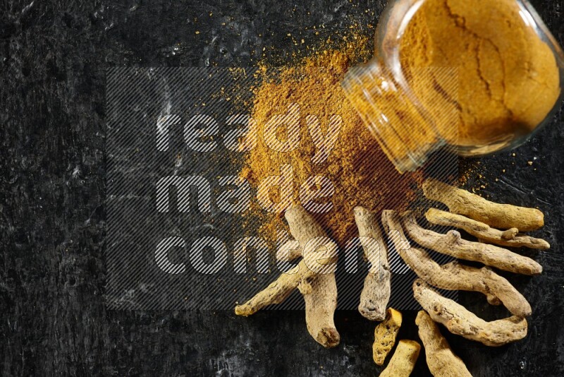 A flipped glass spice jar full of turmeric powder and powder spilled out of it with dried whole fingers on textured black flooring