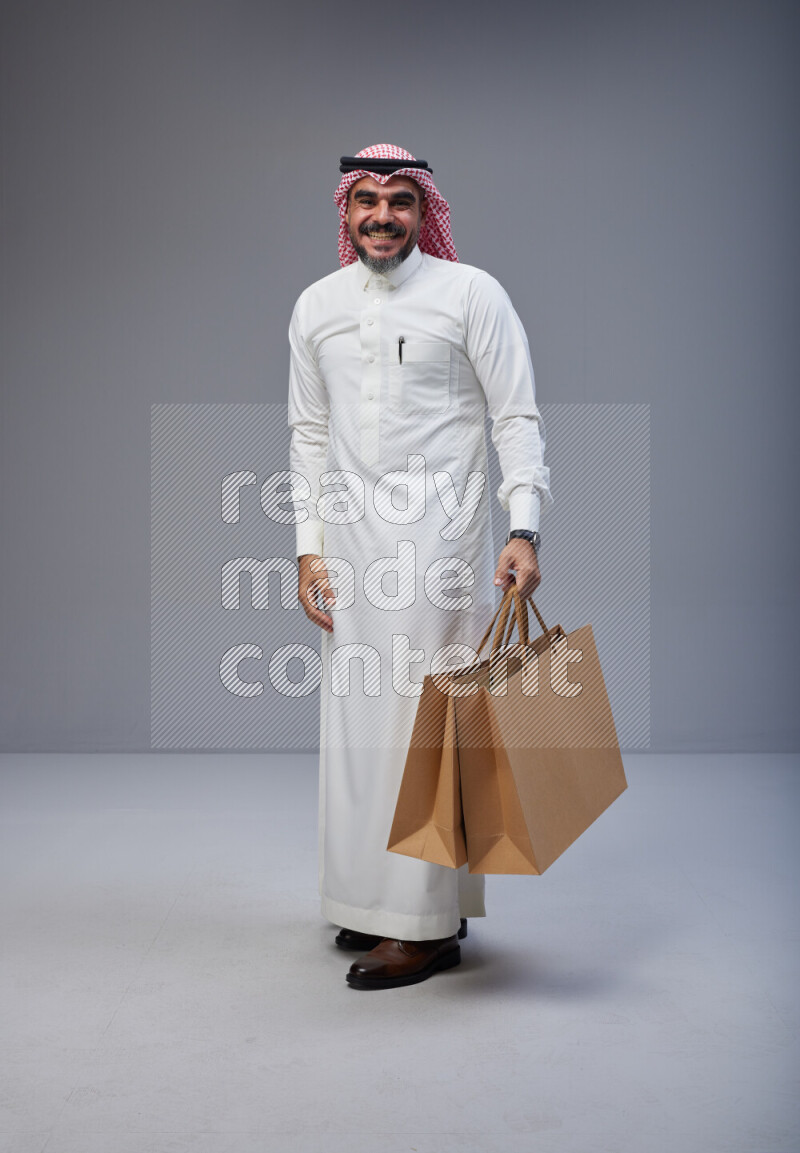 Saudi man Wearing Thob and red Shomag standing holding shopping bag on Gray background