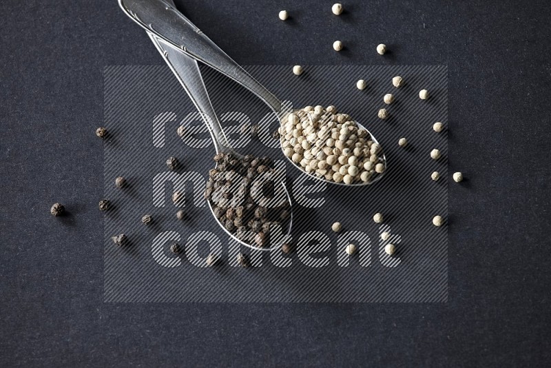 2 metal spoons full of black and white pepper beads with spreaded beads on black flooring