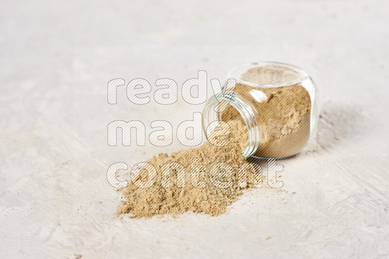 A glass jar full of ground ginger powder flipped with some spilling powder on white background