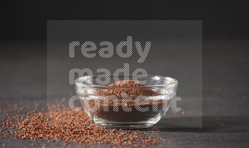 A glass bowl full of garden cress seeds surrounded by seeds on a black flooring
