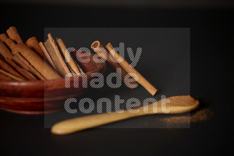 Cinnamon sticks in wooden bowl and cinnamon powder in a wooden spoon on black background