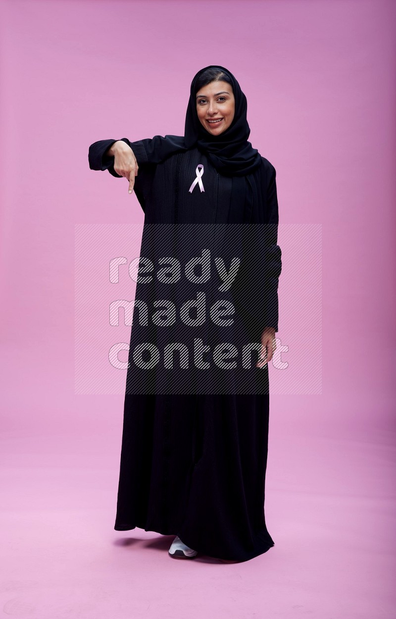 Saudi woman wearing pink ribbon on Abaya standing interacting with the camera on pink background