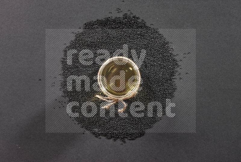 A glass jar full of black seeds oil surrounded by the seeds on a black flooring