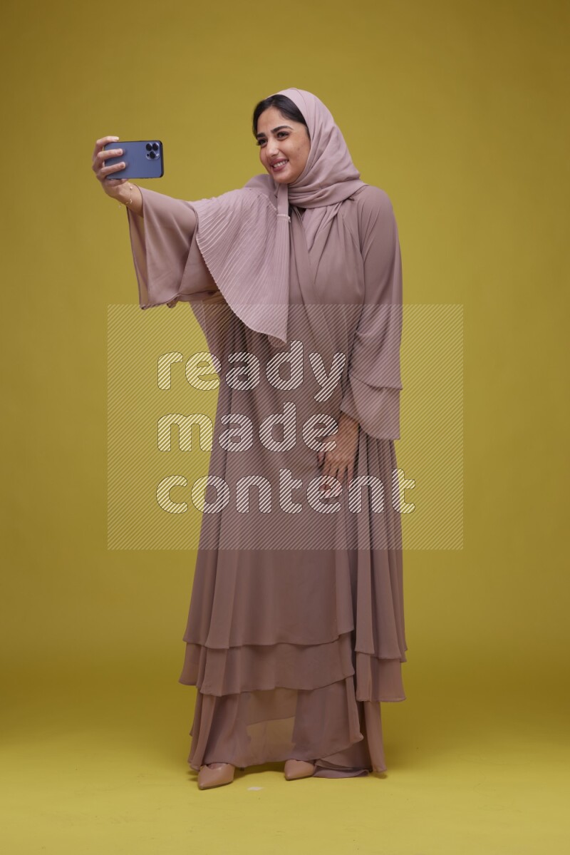 A woman taking a Selfie on a Yellow Background wearing Brown Abaya with Hijab