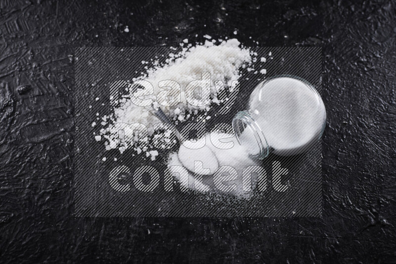 A glass jar full of table salt with some sea salt crystals beside it on a black background