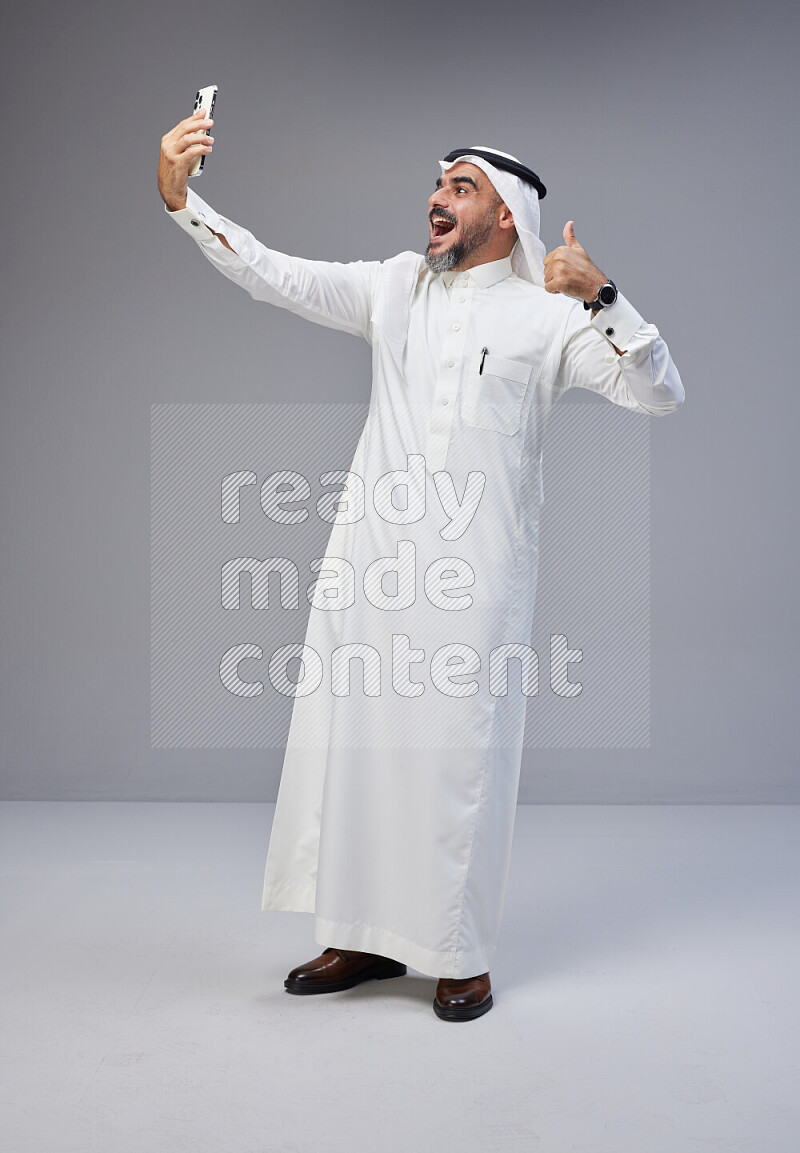 Saudi man Wearing Thob and white Shomag standing taking selfie on Gray background