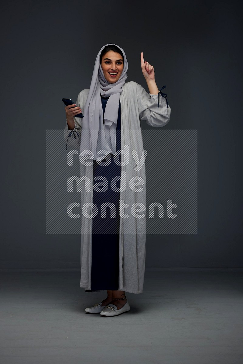 A Saudi woman wearing a light gray Abaya and head scarf standing and holding a phone on a grey background