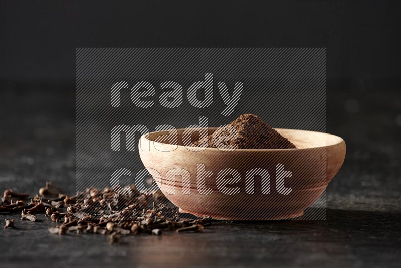 A wooden bowl full of cloves powder on a textured black flooring