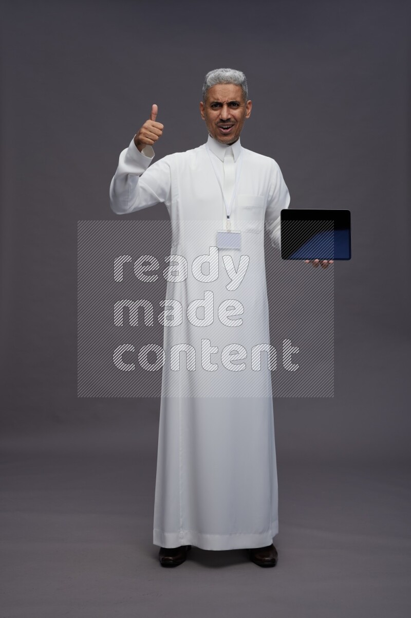 Saudi man wearing thob with neck strap employee badge standing showing tablet to camera on gray background