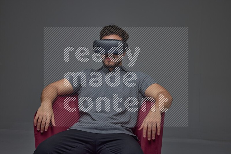 A man wearing casual and wearing VR while sitting on a burgundy chair eye level on a gray background