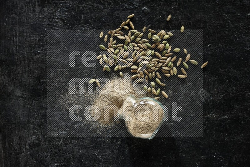 A flipped glass spice jar full of cardamom powder and powder spilled out of it with cardamom seeds spreaded on textured black flooring
