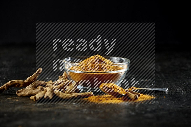A glass bowl and a metal spoon full of turmeric powder and dried whole fingers next of them on textured black flooring