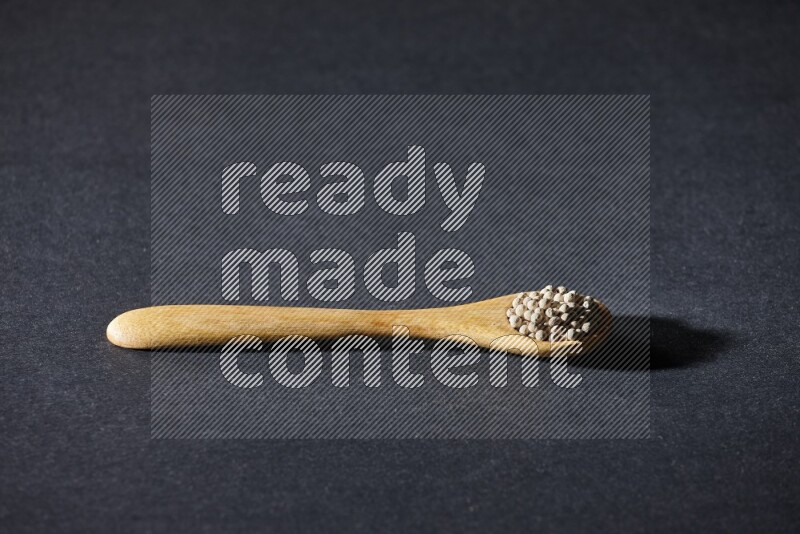 A wooden spoon full of white pepper beads on black flooring
