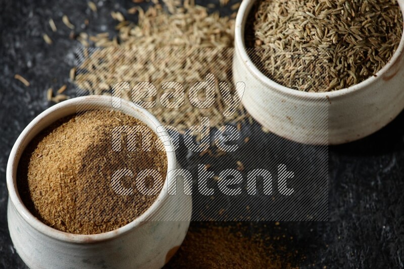 2 beige bowls, one full of cumin powder and the other full of seeds on a textured black flooring