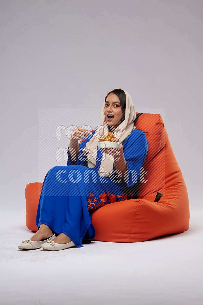 A Woman sitting on an orange beanbag wearing Jalabeya holding a plate of luqaimat