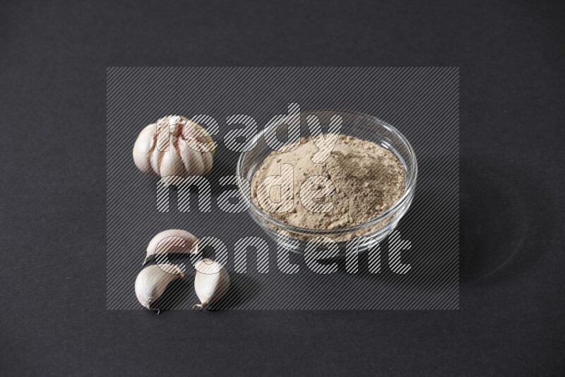 A glass bowl full of garlic powder with garlic bulb and some cloves beside it on a black flooring