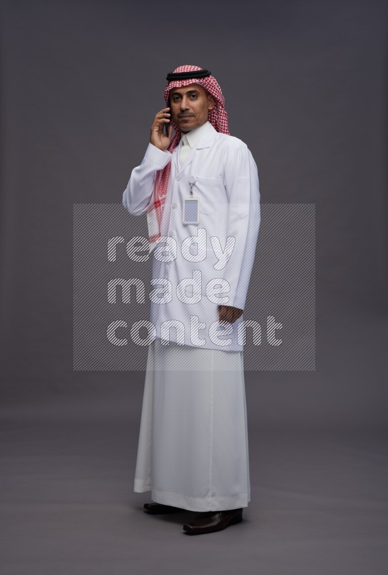 Saudi man wearing thob with lab coat and shomag with pocket employee badge standing talking on phone on gray background