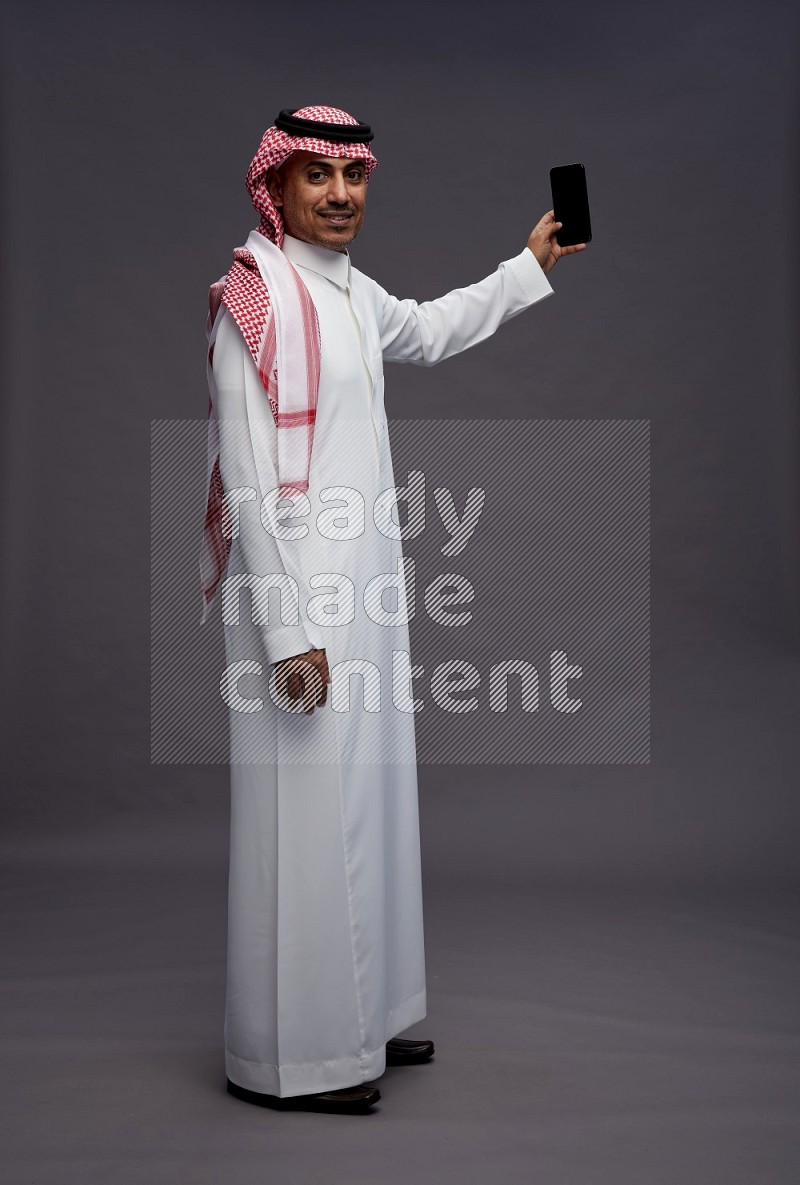 Saudi man wearing thob and shomag standing taking selfie on gray background