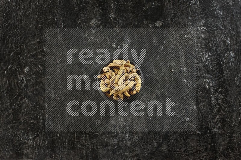 A black pottery bowl full of dried turmeric whole fingers on textured black flooring
