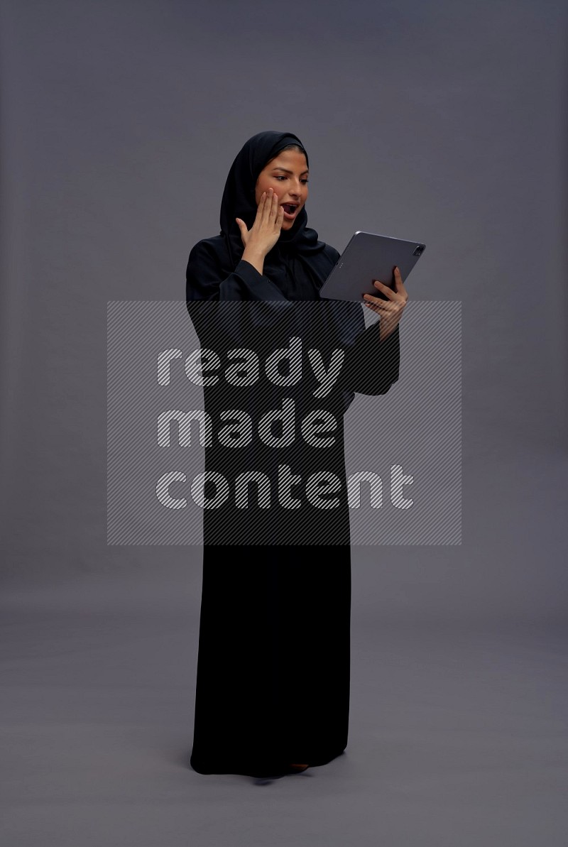 Saudi woman wearing Abaya standing working on tablet on gray background