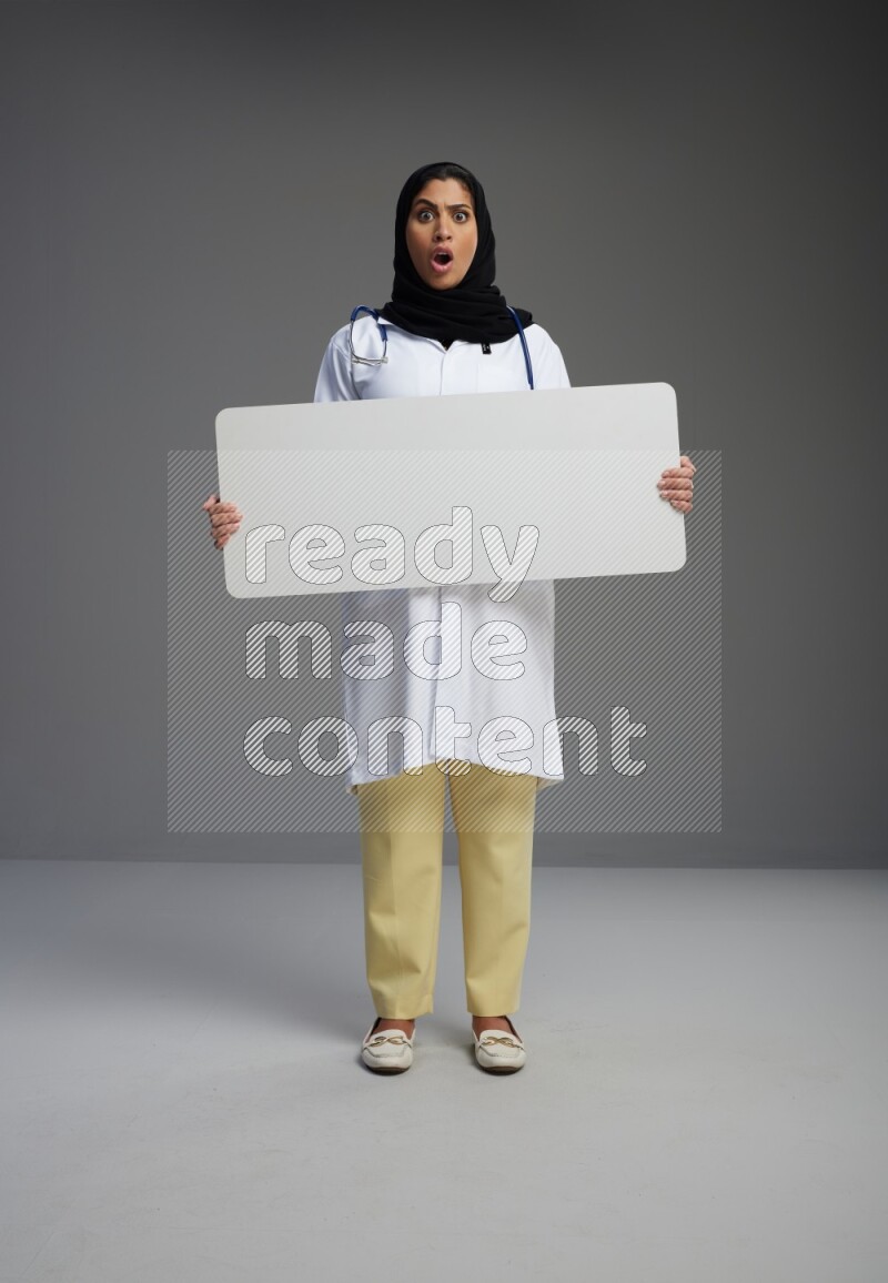 Saudi woman wearing lab coat with stethoscope standing holding board on Gray background