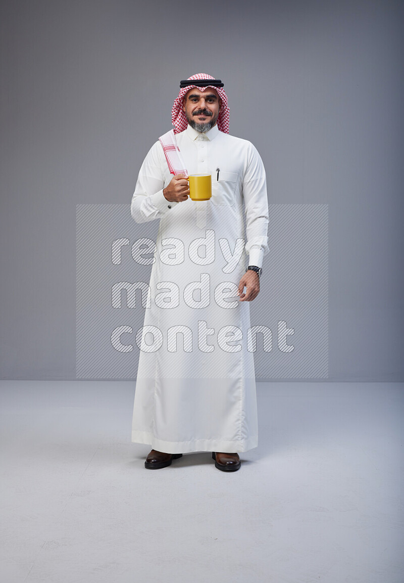 Saudi man Wearing Thob and red Shomag standing holding mug on Gray background