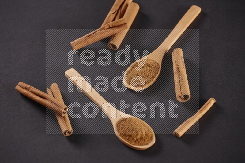 Two wooden spoons full of cinnamon powder with cinnamon sticks on black background