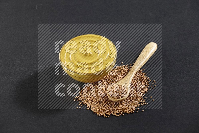 A glass bowl full of mustard paste with mustard seeds underneath and a full wooden spoon on black flooring