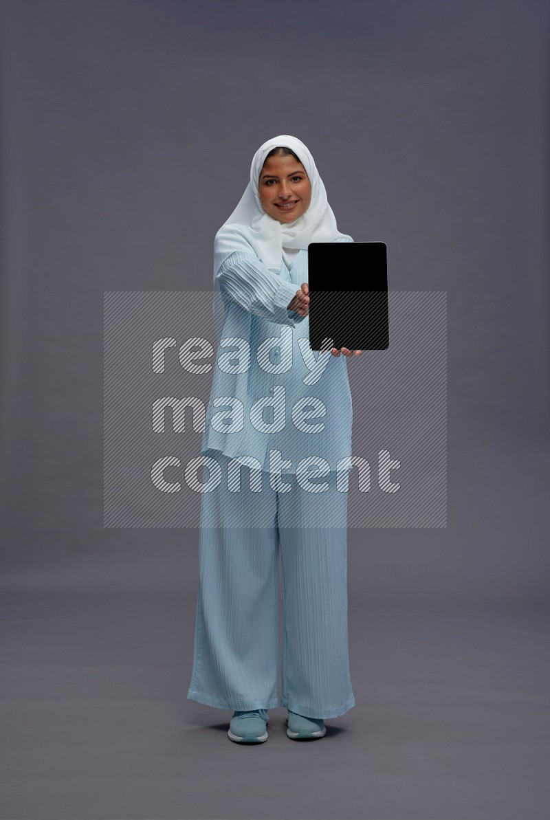 Saudi woman wearing hijab clothes standing showing tablet to camera on gray background