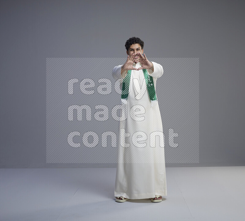 A Saudi man standing wearing thob and Saudi flag scarf on gray background