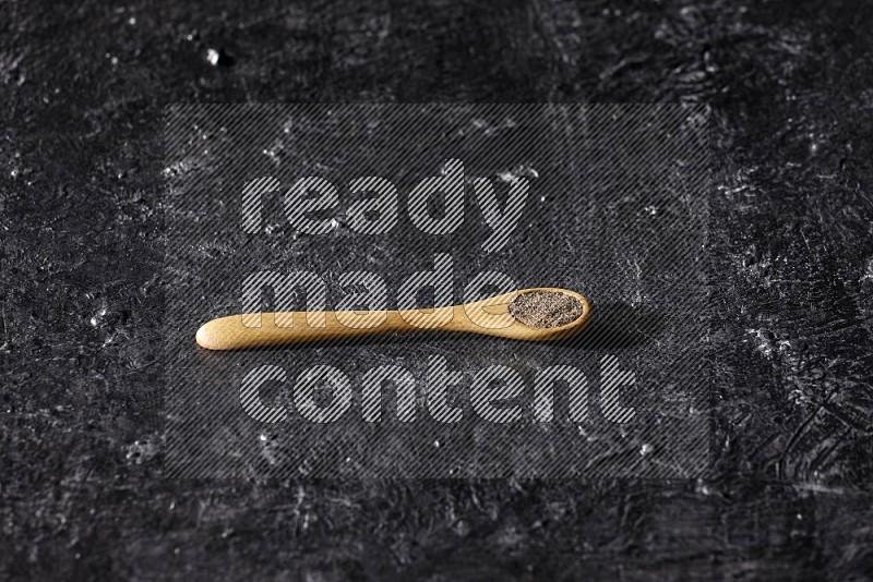 A wooden spoon full of black pepper powder on a textured black flooring