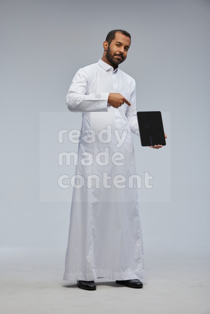 Saudi man wearing Thob standing showing tablet to camera on Gray background