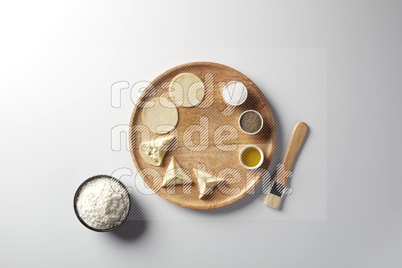 two closed sambosas and one open sambosa filled with cheese while flour, salt, black pepper and oil with oil brush aside in a wooden dish on a white background