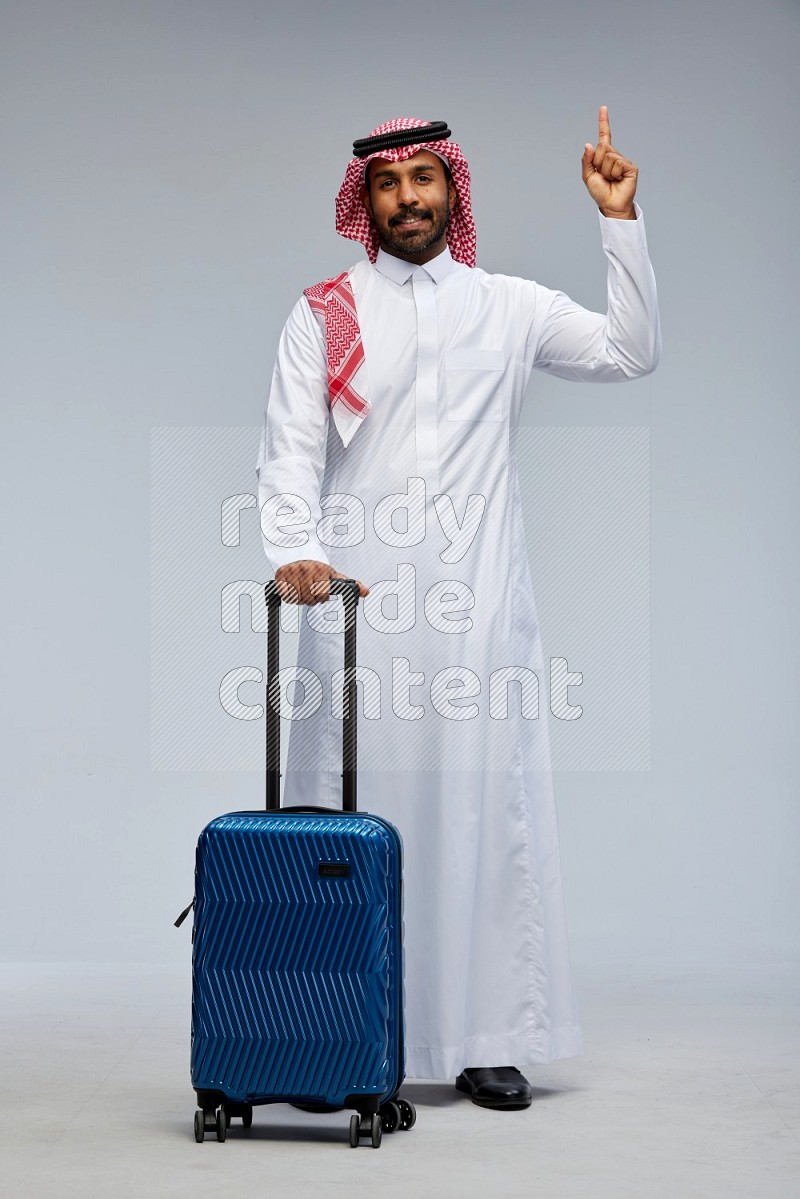 Saudi man wearing Thob and shomag standing holding Travel bag on Gray background
