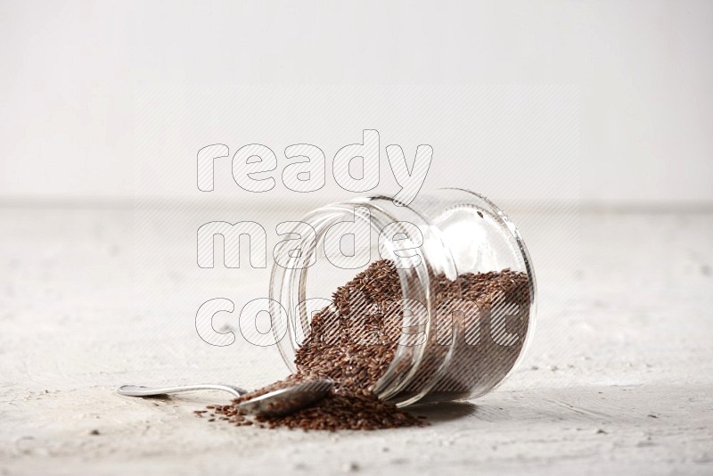 A glass jar full of flax flipped and flax spreaded out with metal spoon full of the seeds on a textured white flooring in different angles