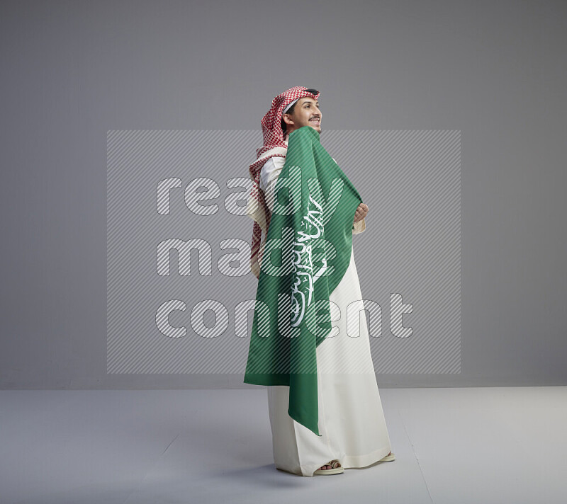 A saudi man standing wearing thob and red shomag holding big saudi flag on gray background