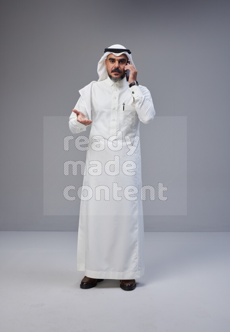 Saudi man Wearing Thob and white Shomag standing talking on phone on Gray background