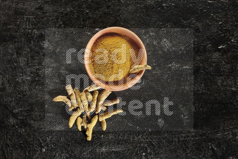A wooden bowl full of turmeric powder with dried turmeric whole fingers on textured black flooring