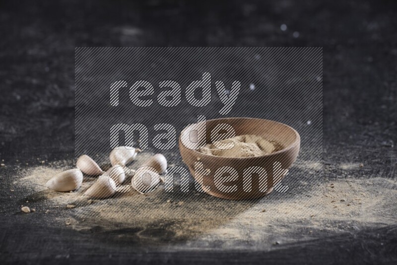 A wooden bowl full of garlic powder with some garlic cloves beside it on a textured black flooring