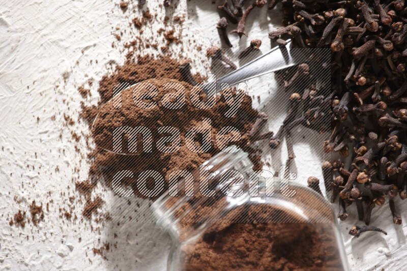 A flipped glass spice jar and a metal spoon full of cloves powder and powder came out of the jar with cloves spread on textured white flooring