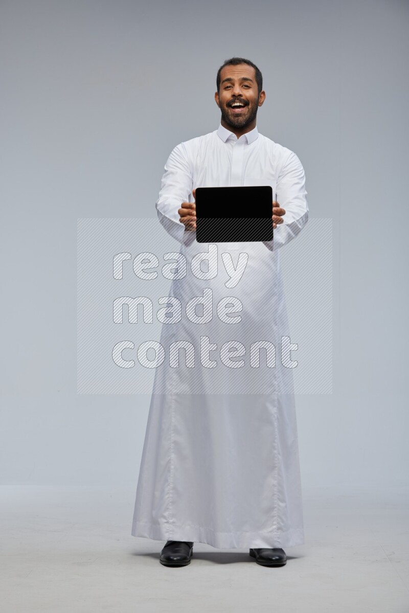 Saudi man wearing Thob standing showing tablet to camera on Gray background