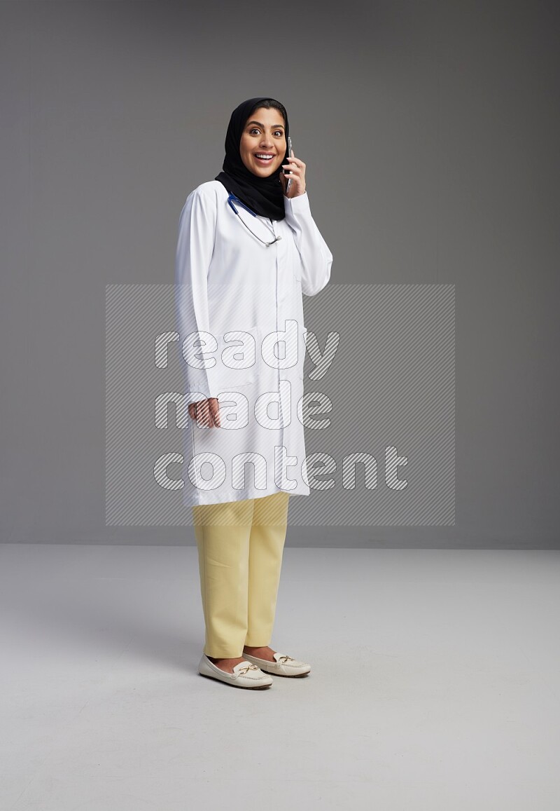 Saudi woman wearing lab coat with stethoscope standing talking on phone on Gray background