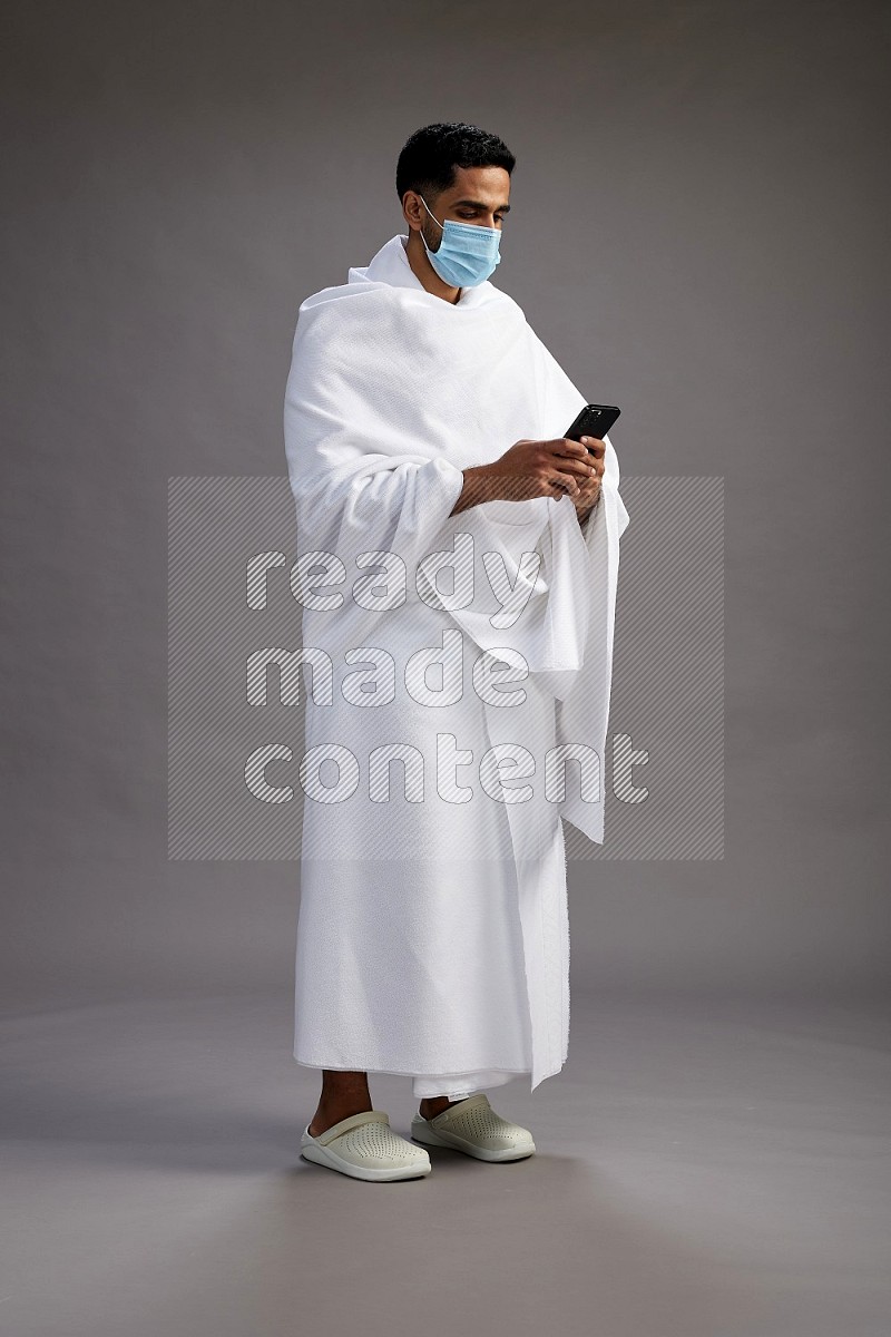 A man wearing Ehram with face mask Standing texting on phone on gray background