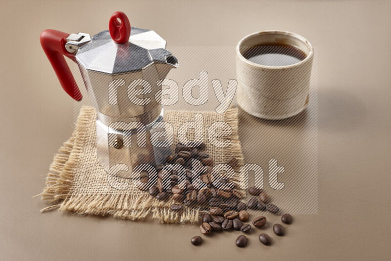 A moka pot with red handle surrounded by roasted coffee beans on beige background