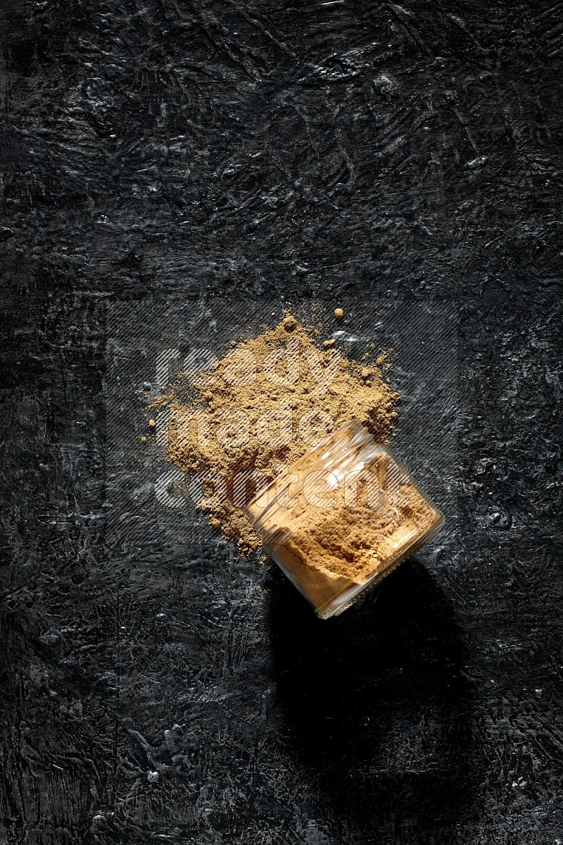 A flipped glass jar full of allspice powder and powder spilled out of it on a textured black flooring