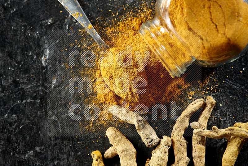 A flipped glass spice jar and metal spoon full of turmeric powder and powder spilled out of it with dried whole fingers on textured black flooring